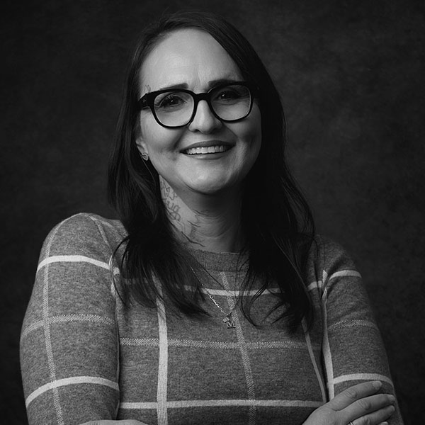 Black and white portrait of a smiling woman with long dark hair, wearing glasses and a patterned sweater. She has a visible tattoo on her neck and is posing with her arms crossed against a dark background.