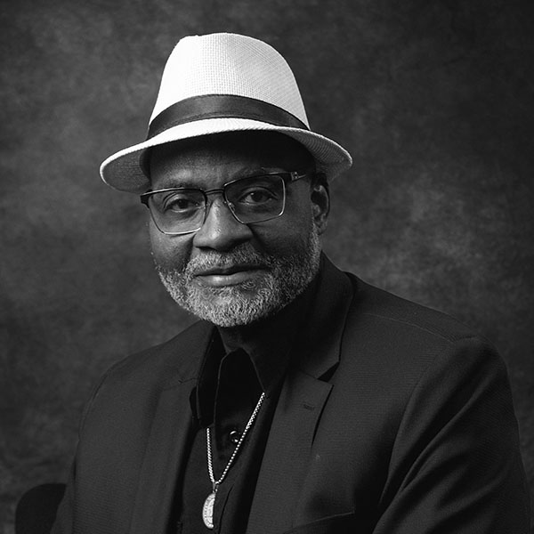 Black and white portrait of an older man wearing glasses, a white fedora, a dark blazer, and a necklace. He has a short gray beard and is smiling softly, facing the camera against a textured background.