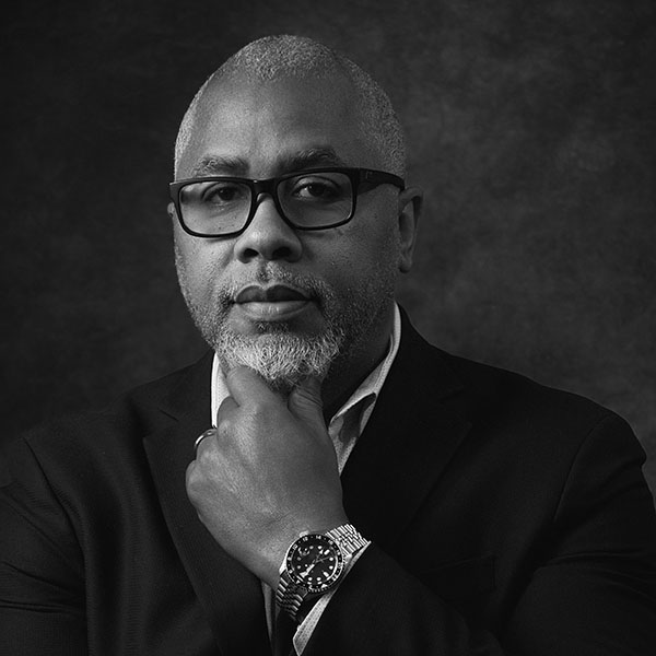 Black and white portrait of a man with gray hair and beard, wearing glasses, a suit, and a wristwatch. He is looking at the camera with one hand touching his chin thoughtfully against a dark background.