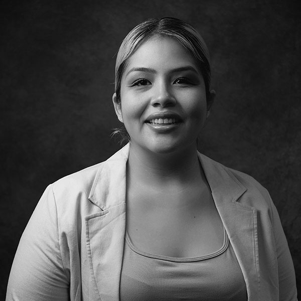 Black and white portrait of a woman smiling, wearing a light-colored blazer over a top, with her hair pulled back, standing against a dark, textured background.