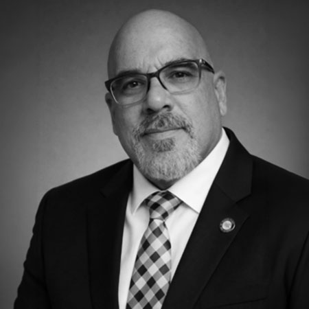Black-and-white portrait of a man with glasses, a bald head, and a goatee, wearing a suit, white shirt, and a checkered tie, posing against a plain background.