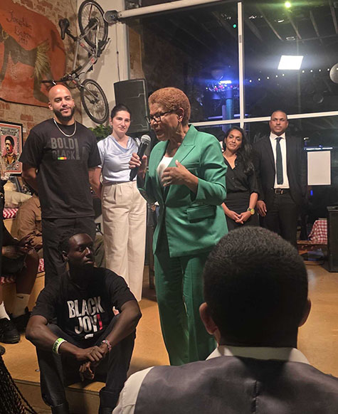 A woman in a green suit speaks into a microphone to a seated audience, demonstrating the importance of civic engagement. Several people stand behind her, some smiling. The setting appears to be an indoor event with both casual and business attire, reflecting a community gathering focused on participation and public involvement.