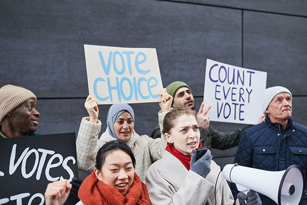 A diverse group of people protesting with signs that read VOTE CHOICE and COUNT EVERY VOTE while one person speaks into a megaphone.